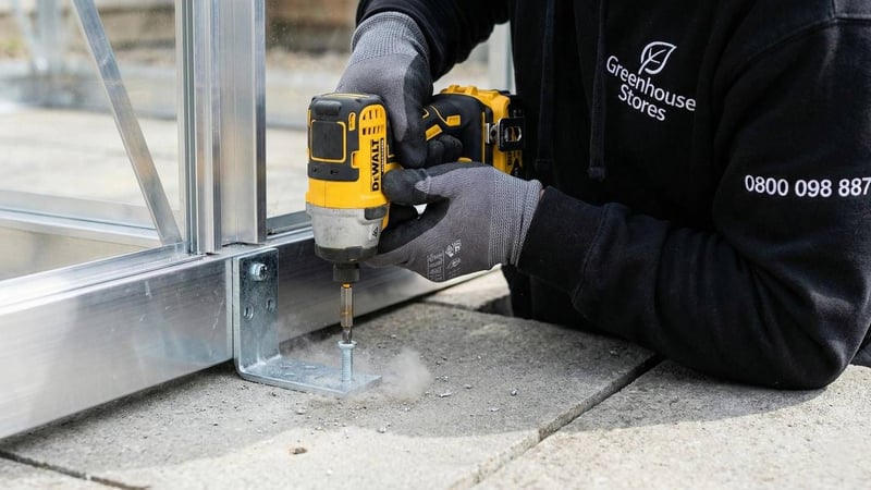 Professional installer bolting greenhouse base bracket into concrete paving slabs to prevent storm damage.