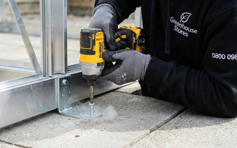 Professional installer bolting greenhouse base bracket into concrete paving slabs to prevent storm damage.