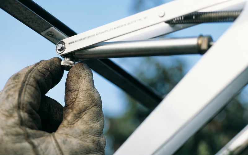 Gloved hand turning the small silver adjustment knob on the wax cylinder of a Bayliss MK7 automatic vent opener fitted to a UK greenhouse