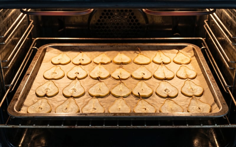 Pear half-slices on a baking tray lined with greaseproof paper inside a domestic oven at low temperature, evenly spaced for drying