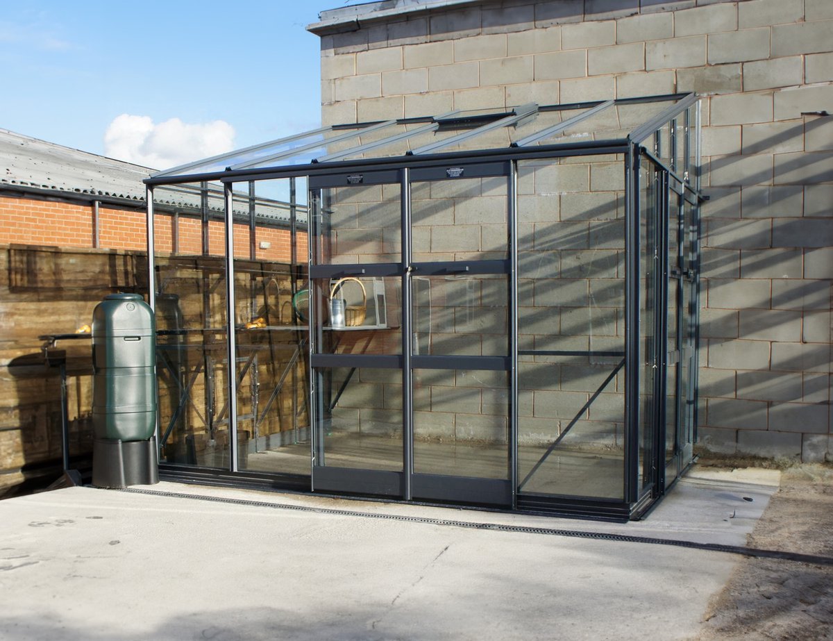 Elite Titan K800 8x12 lean-to greenhouse with heavy-gauge Titan box-section aluminium glazing bars and toughened glass, fitted against a UK brick wall on a concrete foundation — the only mainstream UK lean-to built with the same frame gauge as the freestanding Titan range